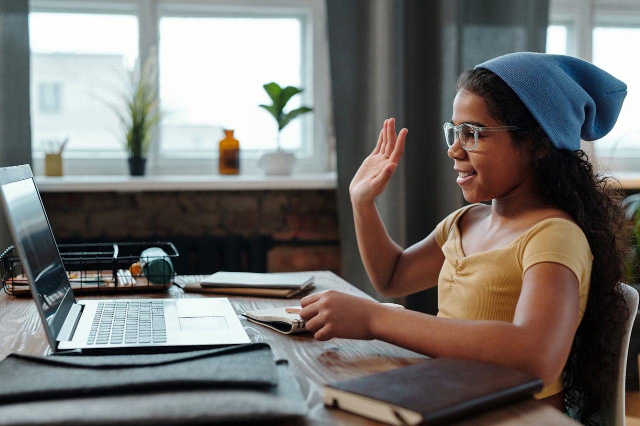 Accueil A young girl waves at her laptop during a video call in a cozy home office setting.